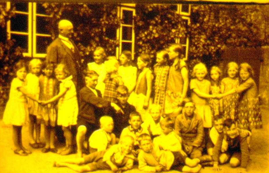 1924 Schulfoto Schmalensee: Das Bild zeigt eine gr��ere Gruppe von Sch�lerinnen und Sch�lern, die im Freien vor dem Schmalenseeer Schulgeb�ude posiert. Die Aufnahme stammt aus dem Jahr 1924 und ist in einem warmen, leicht vergilbten Ton gehalten. Die Kind